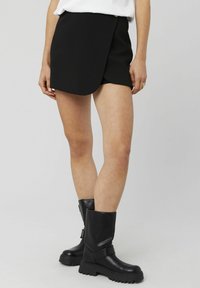 Mini jupe noire en tissu lisse, avec un détail de bouton sur le côté. Assortie à des bottes en cuir noires hautes jusqu'aux genoux avec une semelle épaisse.