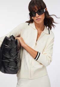 Maglione crema con zip con strisce nere al collo e ai polsi, abbinato a una borsa a sacchetto nera lucida tenuta sotto il braccio sinistro.