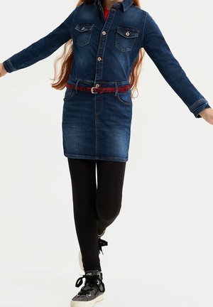 Barn iført en blå denim kjole med knapper, et rødt bælte, sorte leggings og sorte sneakers med hvide såler, stående med armene spredt.