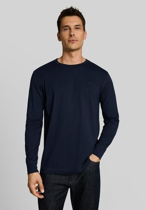 Man draagt een marineblauw overhemd met lange mouwen en donkere spijkerbroek, staand met één hand in de zak tegen een effen grijze achtergrond.