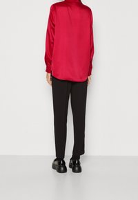 Czerwona satynowa koszula z długim rękawem i zaokrąglonym dołem, w zestawie z czarnymi spodniami i błyszczącymi czarnymi butami. Materiał ma gładkie, lśniące wykończenie.