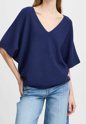 Kvinde iført en løs mørkeblå V-hals-top med brede korte ærmer og lyseblå cowboybukser, poserer med den ene hånd på hoften.