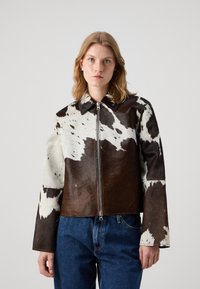 Braun-weiße Kuhhaut-Lederjacke mit Reißverschluss, spitzem Kragen und weiten Ärmeln, kombiniert mit blauen Jeans.