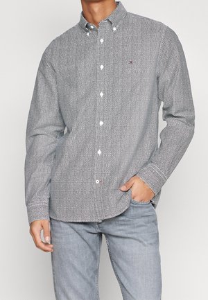 Homme portant une chemise à manches longues à carreaux noirs et blancs boutonnée, un jean gris clair, une main dans la poche, debout devant un fond uni.