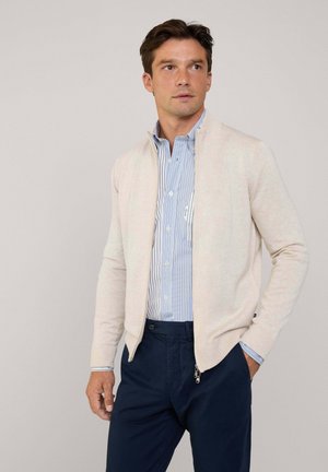 Light beige zip-up cardigan, lightweight knit texture, worn over a blue striped dress shirt, paired with dark navy trousers.