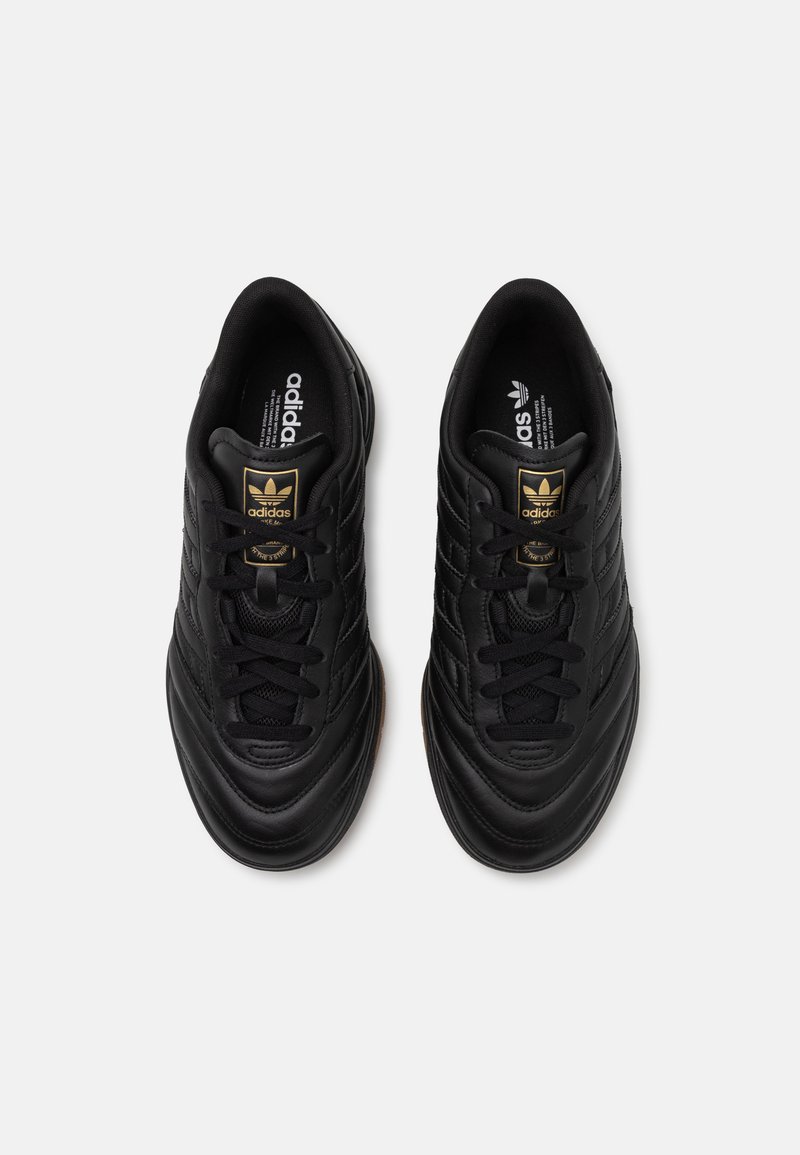 Zapatillas deportivas negras con una parte superior de cuero liso, costuras texturizadas, cordones negros y un logotipo dorado en la lengüeta. Suela de goma para agarre.