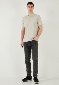 Helle beige Polohemden mit einem Reißverschlusskragen, kombiniert mit dunkelgrauen Jeans und schwarzen Sneakers, vor einem neutralen Hintergrund stehend.