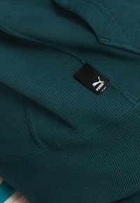 Sudadera color azul verdoso con textura acanalada, que presenta una pequeña etiqueta negra con el logo de Puma cosida en la tela. Material suave y diseño casual.
