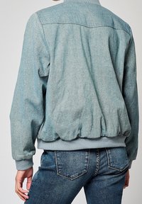 Veste bomber bleu clair en tissu texturé, avec poignets côtelés et ourlet froncé, dotée d'un col rond simple et d'un détail au plastron arrière.