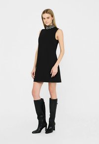 Vestido negro sin mangas con cuello alto y collar adornado, combinado con botas negras de cuero hasta la rodilla, creando una silueta minimalista.