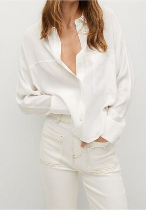 Žena v volné bílé košili na knoflíky a vysokým pasem bílých kalhotách s viditelnými předními kapsami, ruce částečně v kapsách.