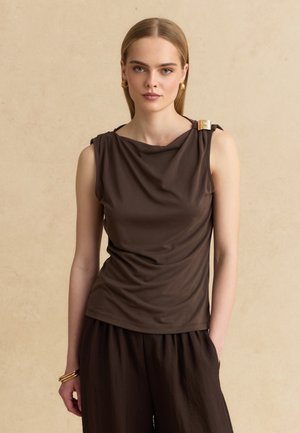Giovane donna con capelli biondi lisci indossa un top senza maniche marrone scuro con dettagli dorati e pantaloni larghi abbinati, posando contro un muro beige.