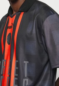 Jersey negro con llamativas rayas verticales rojas y un patrón de cuadros texturizado. Incluye un collar negro y detalles del logotipo.