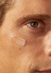 Nahaufnahme eines Gesichts mit hellbrauner Haut, einem markanten braunen Auge und einem durchscheinenden, gelartigen Produkt, das auf die Wange aufgetragen wurde.