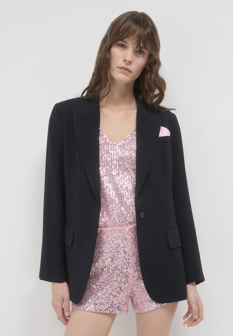 Blazer nero con vestibilità sartoriale, rever a scendere e tasche frontali, indossato sopra un top di paillettes rosa e pantaloni corti abbinati.