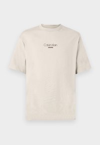 Bežs īsām piedurknēm T-krekls ar "Calvin Klein JEANS" logotipu centrā uz krūtīm.