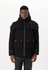 Chaqueta negra con capucha, con epaulets con tachuelas y bolsillos con botones a presión. Hecha de tejido texturizado con cremallera frontal.