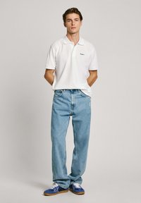 Fehér póló, világoskék farmernadrág és kék sportcipő. A pólón kis logó található a mellrészen. Az outfit laza, kényelmes fazonú.