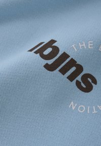 Ljusblå texturerad tyg med en framträdande mörk svart logotyp som överlappar texten "THE ORIGINAL" i vitt, vilket skapar en lagerdesigneffekt.