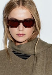 Zonnebril met een donkerbruine, oversized, geometrische frame. De glazen zijn getint in bruin. Het model draagt een bruine gebreide trui met een kraag.