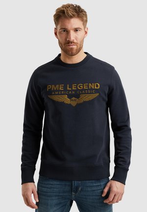 Námornícky sveter s okrúhlym výstrihom, ktorý má zlatý nápis "PME LEGEND AMERICAN CLASSIC" a grafiku krídiel. Mäkká textúra, rebrované manžety a lem.