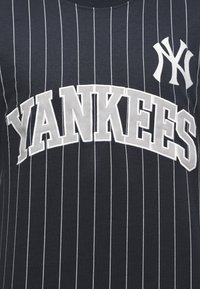 Πουκάμισο σκούρου ναυτικού χρώματος με λεπτές ρίγες, μεγάλο λευκό κείμενο «YANKEES» και το λογότυπο των New York Yankees στο πάνω δεξί στήθος.