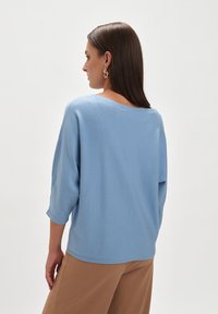 Jasnoniebieski, oversize sweter z rękawami o długości trzech czwartych, o miękkiej fakturze i szerokim dekolcie, w parze z beżowymi spodniami o szerokich nogawkach.