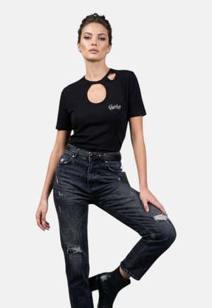 Donna in piedi con una gamba sollevata, indossa una t-shirt nera con ritagli, jeans neri strappati a vita alta e stivaletti neri su uno sfondo semplice.