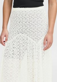Jupe maxi en dentelle blanche avec motif floral, présentant un design superposé, une taille élastique et une texture transparente. Main visible à la taille.
