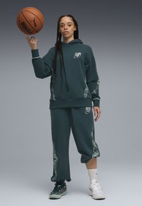 Sudadera de color verde oscuro y joggers a juego con patrones decorativos en verde claro y logo blanco. El modelo sostiene una pelota de baloncesto, vistiendo zapatillas bicolor.