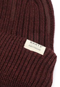 Bordeauxrode geribde gebreide muts met een opgevouwen boord, voorzien van een rechthoekig crèmekleurig label met de tekst "BARTS" en "AMSTERDAM" in het zwart gedrukt.