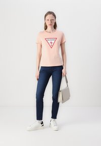 Tricou din bumbac de culoare roz deschis, cu un design grafic triunghiular roșu care prezintă cuvântul "GUESS" și un semn de întrebare, asociat cu blugi de culoare albastră închis și adidași albi.