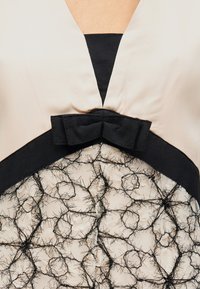 Μπεζ φόρεμα με μαύρη V-λαιμόκοψη, μαύρο υφασμάτινο φιόγκο στη μέση και διάφανη δαντελένια φούστα με ακανόνιστα μαύρα νήματα.