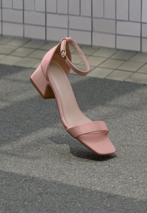 Single pink block-heeled sandal with ankle strap resting on asphalt near tiled wall background.
