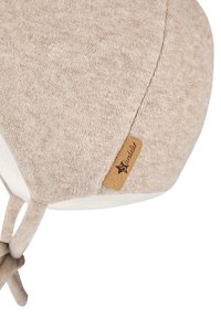 Beige fleece hat with a soft texture, curved shape, and a fabric tag featuring the brand name. Drawstring accents for adjustments.