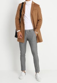 Ein brauner Wollmantel über einem weißen Rollkragenpullover, graue schmal geschnittene Hose und weiße Sneaker, hält eine schwarze Umhängetasche. Einfaches Design, maßgeschneiderte Passform.