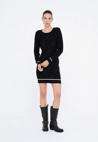 Robe noire en maille à manches longues, avec des lignes blanches diagonales en accent et un ourlet rayé blanc. Associée à des bottes noires montantes jusqu'aux genoux.
