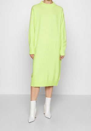 Knit dress in bright neon green, featuring a high neckline and long sleeves, paired with glossy white ankle boots and textured ribbed cuffs.