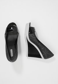 Svarta kilklackar med en glänsande finish och subtil märkning, som visar en öppen tå-design och en kontrasterande sidopanel.