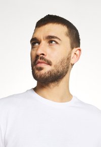 White cotton t-shirt with a round neckline, minimal texture, worn by a man with short brown hair and light stubble, against a light background.