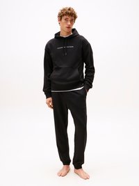 Felpa nera con tasca frontale e branding bianco, abbinata a pantaloni sportivi neri con stampa del logo laterale. Tessuto morbido, vestibilità rilassata.