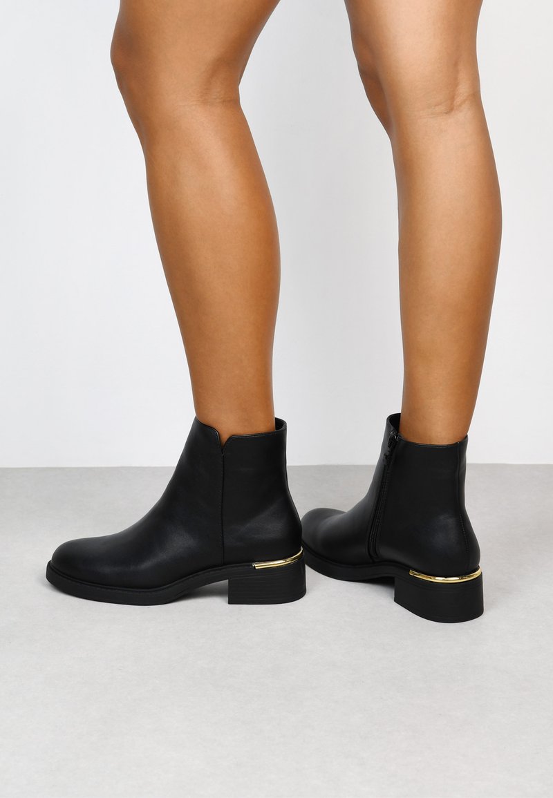 Botas de cuero negras hasta el tobillo con un tacón bajo en bloque, con un acento dorado y cierre con cremallera lateral. Textura suave, diseño de punta redondeada.