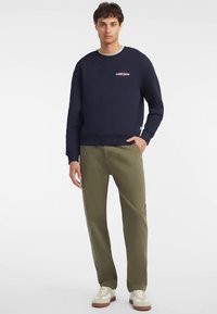 Tengerészkék pulóver kerek nyakkivágással, kis piros logóval a mellkasán. Párosítva olívazöld nadrággal és bézs tornacipővel.