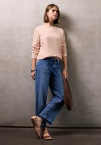 Jeune femme en pull rose clair et jeans large se tient contre un mur en béton, tenant un sac marron, portant des ballerines en bronze.