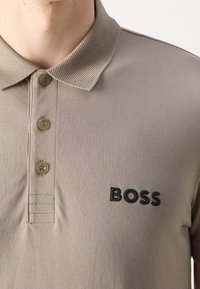 Polo beige avec trois boutons et col texturé, arborant un logo "BOSS" noir brodé sur le côté gauche de la poitrine.