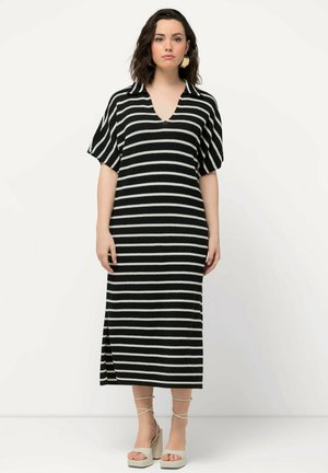 Robe rayée noir et blanc avec des manches courtes, décolleté en V, fente latérale et longueur genou. Le modèle porte des chaussures à plateforme de couleur claire.