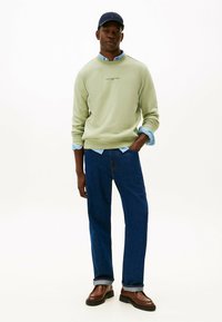Felpa verde chiaro con logo ricamato, indossata sopra una camicia blu a colletto, abbinata a jeans blu scuro e scarpe marroni.