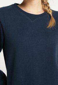 Sudadera azul marino oscura con un diseño de cuello en V, puños acanalados y un sutil logo bordado en el pecho. Fabricada con una tela suave y lisa.