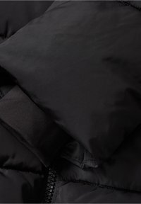 Czarna kurtka puchowa z gładkim materiałem, z zamkiem błyskawicznym i detalem w postaci ściągaczy na mankietach. Całościowy design jest lekki i kompaktowy.