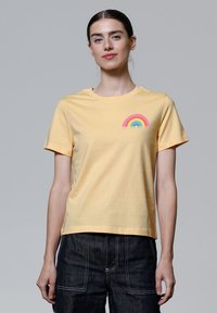 Geel katoenen T-shirt met korte mouwen, ronde halslijn en een kleine regenboogafbeelding in roze, blauw en groen gestikt op de linkerborst.